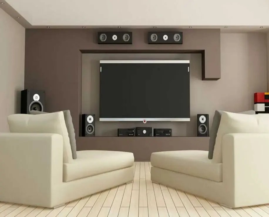 A modern living room featuring two light-colored sofas facing a wall-mounted flat-screen TV, surrounded by speakers and a minimalistic design.