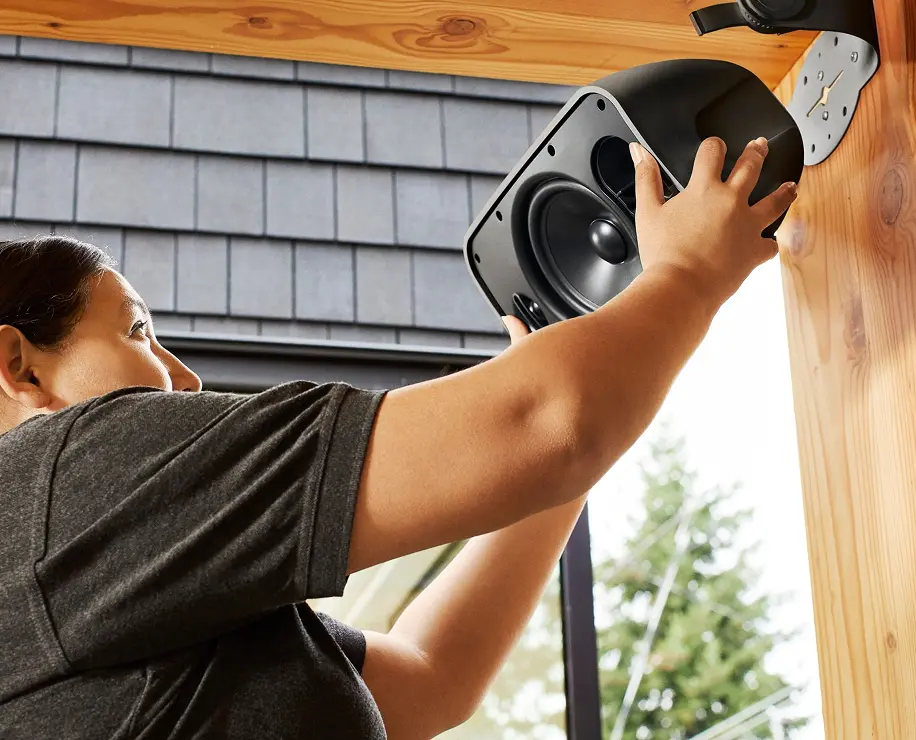A person installing a black speaker onto a wall in a well-lit room with wooden accents.