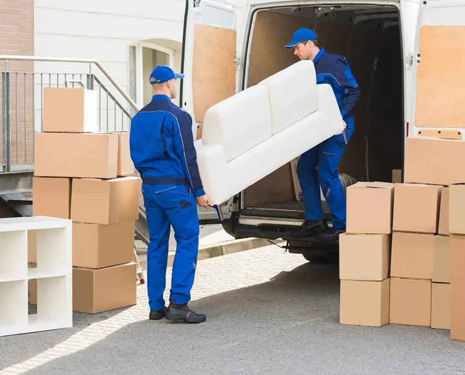 Two movers in blue uniforms are carrying a white sofa out of a moving van, surrounded by cardboard boxes.