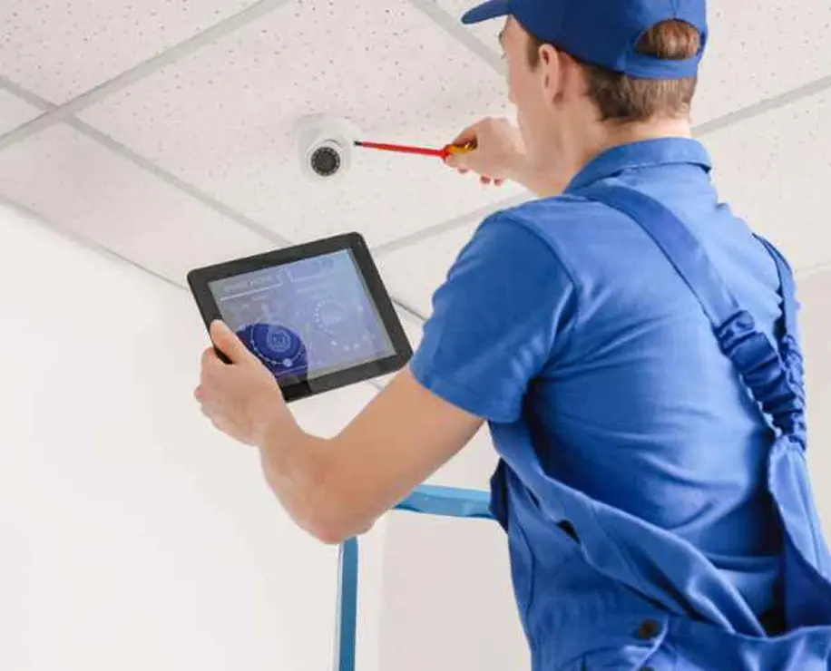 A technician in a blue uniform using a tablet while working on a ceiling-mounted device.