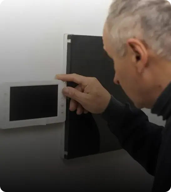 A person interacting with a wall-mounted device, pointing at a screen while standing next to a black panel.