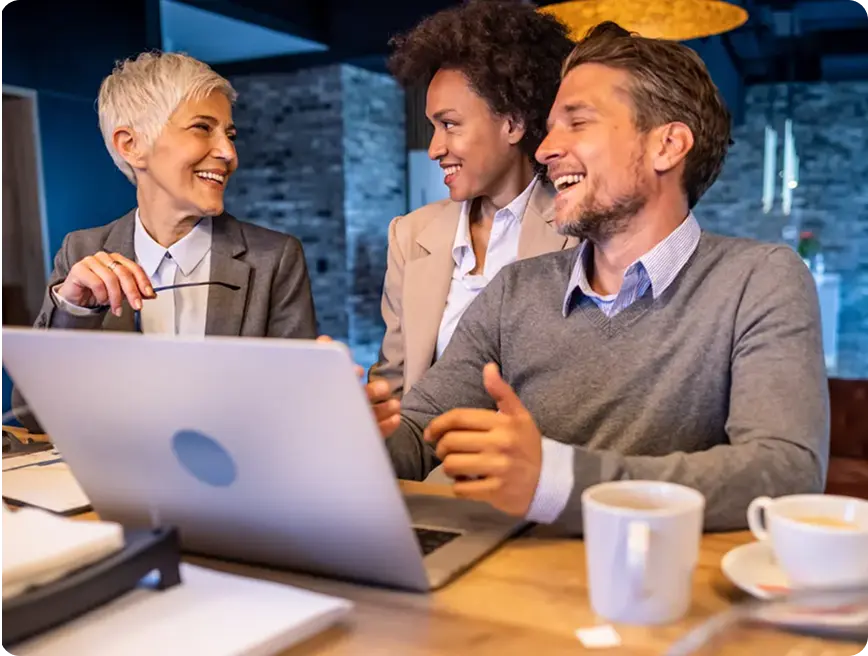 Three professionals engaged in a friendly discussion at a workspace, with a laptop in front of them.