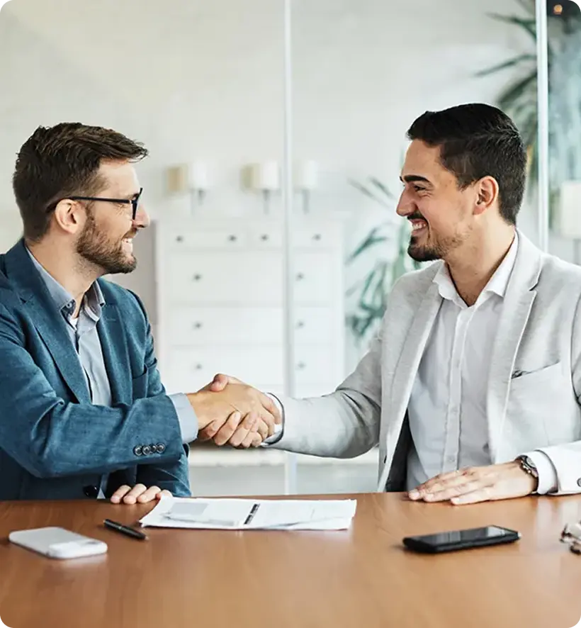 Two men shaking hands across a conference table, smiling, with documents and a phone on the table.