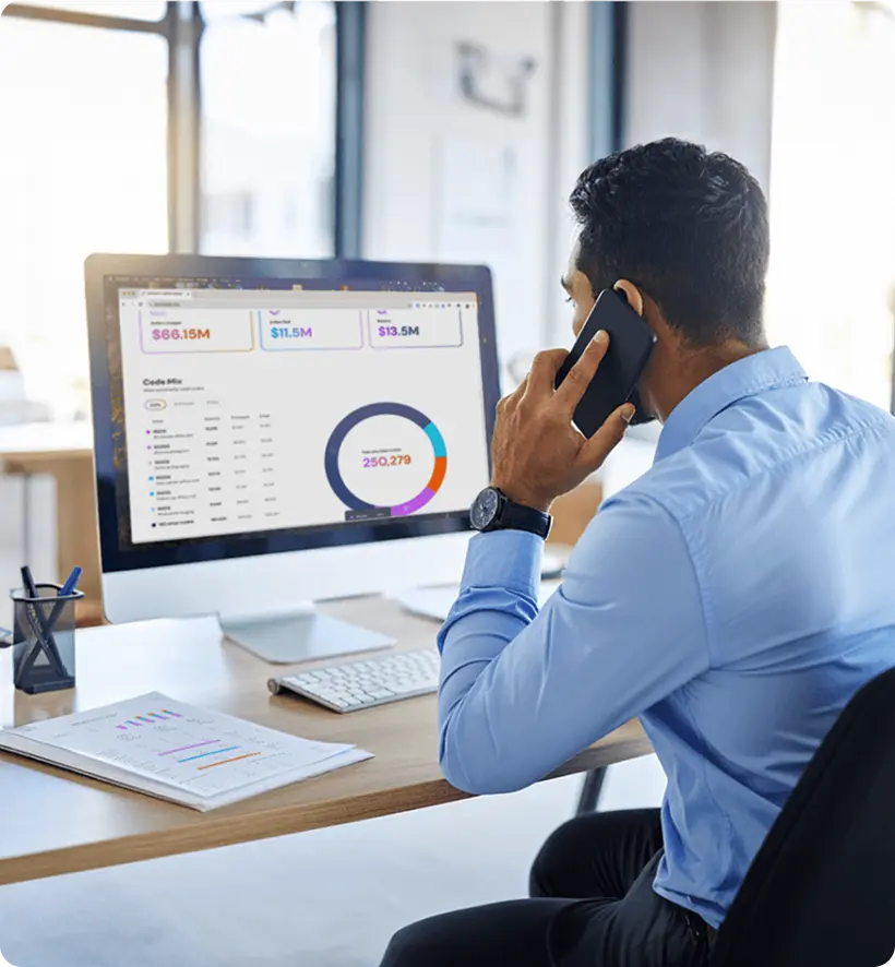 A man in a blue shirt sitting at a desk, talking on the phone while looking at a computer screen displaying financial data and graphs.