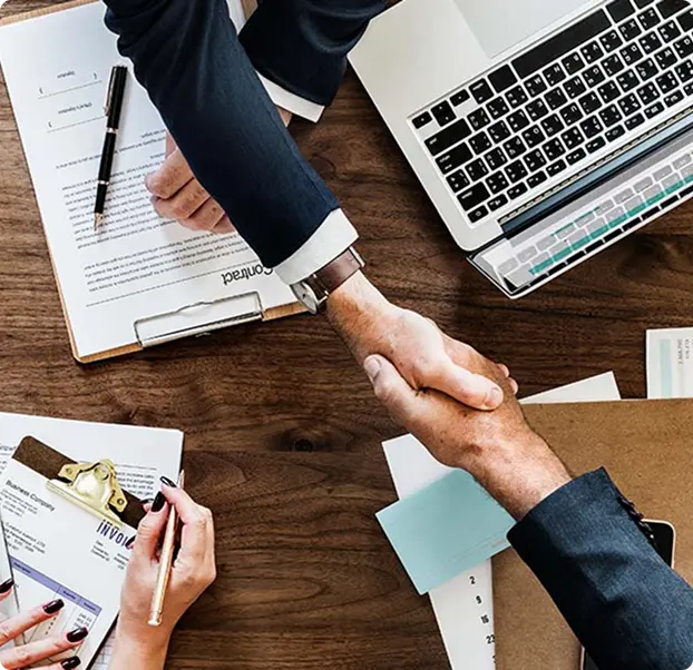 Two hands shaking over a wooden table with documents and a laptop in the background.