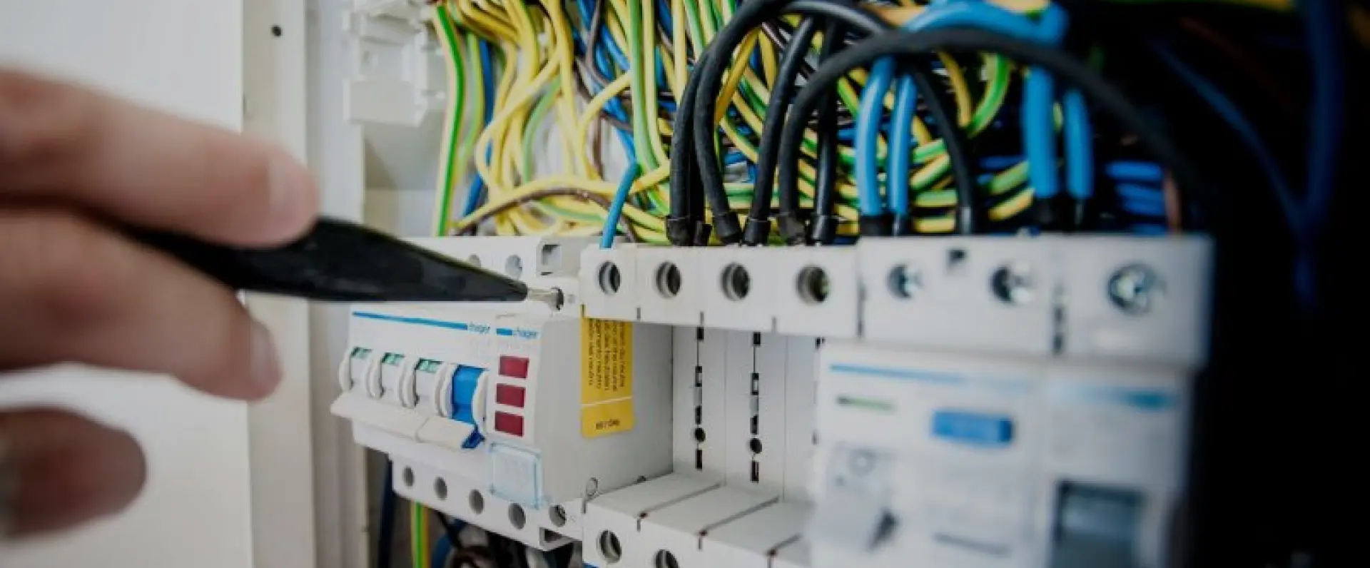 A close-up of a person's hand using a tool on electrical circuit breakers and wiring in a control panel.