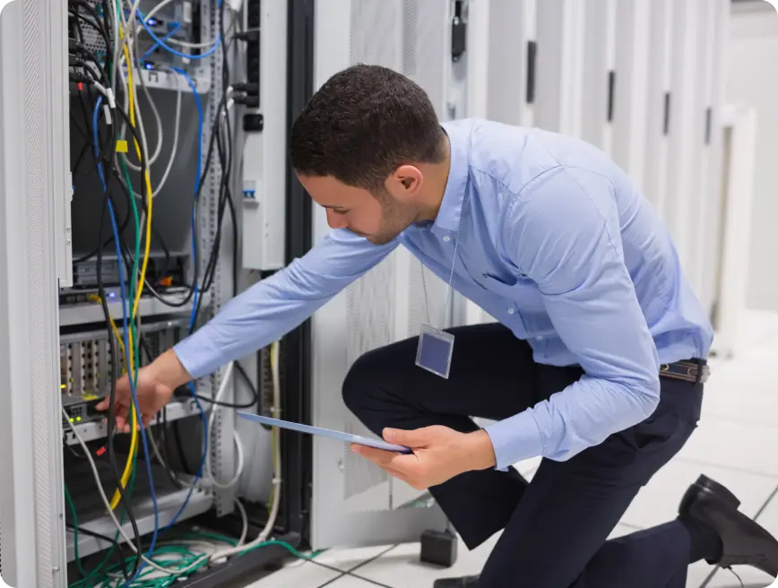 A technician in a blue shirt kneels to check connections in a server room filled with racks of equipment and colorful cables.