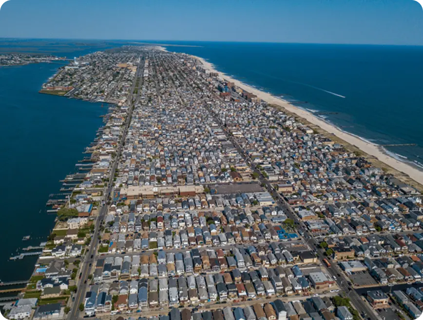 Aerial view of a coastal town with densely packed houses extending towards the beach, with a clear blue ocean on one side and a calm bay on the other.