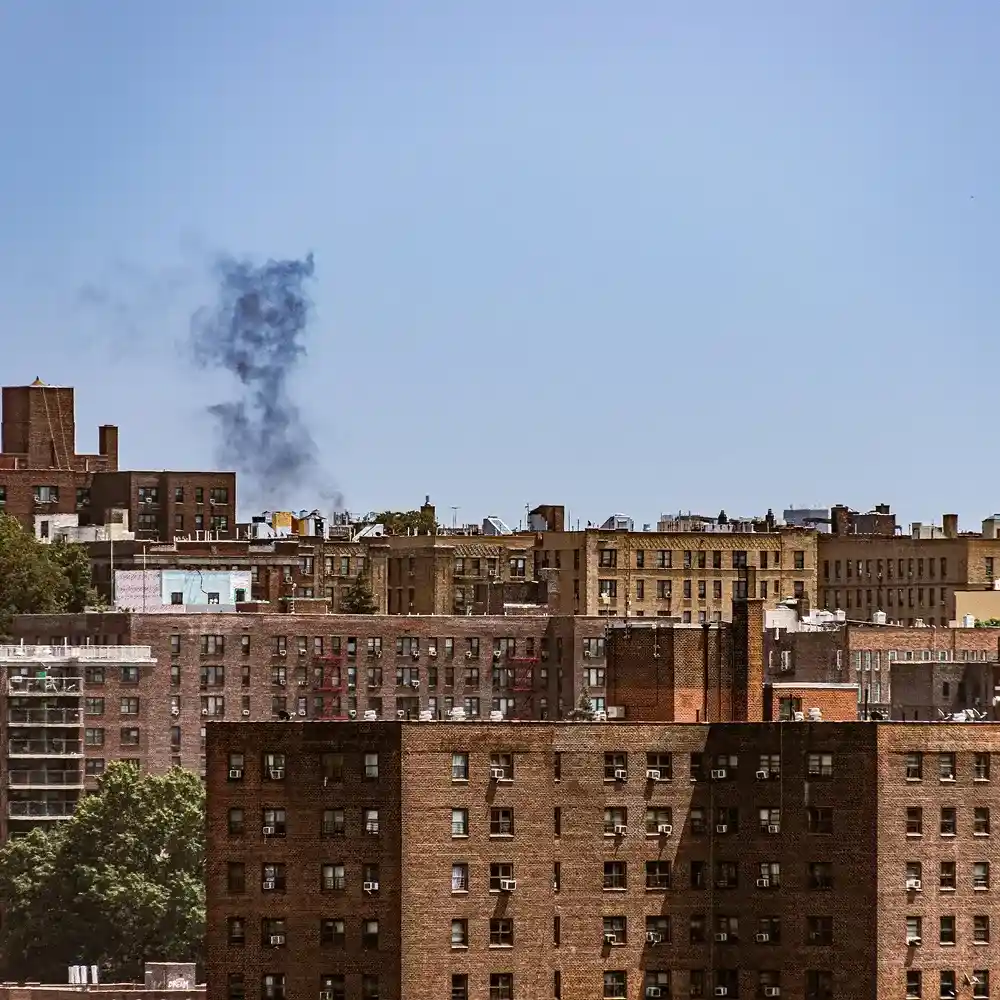 A cityscape featuring multiple buildings with a smoky plume rising in the background against a clear blue sky.
