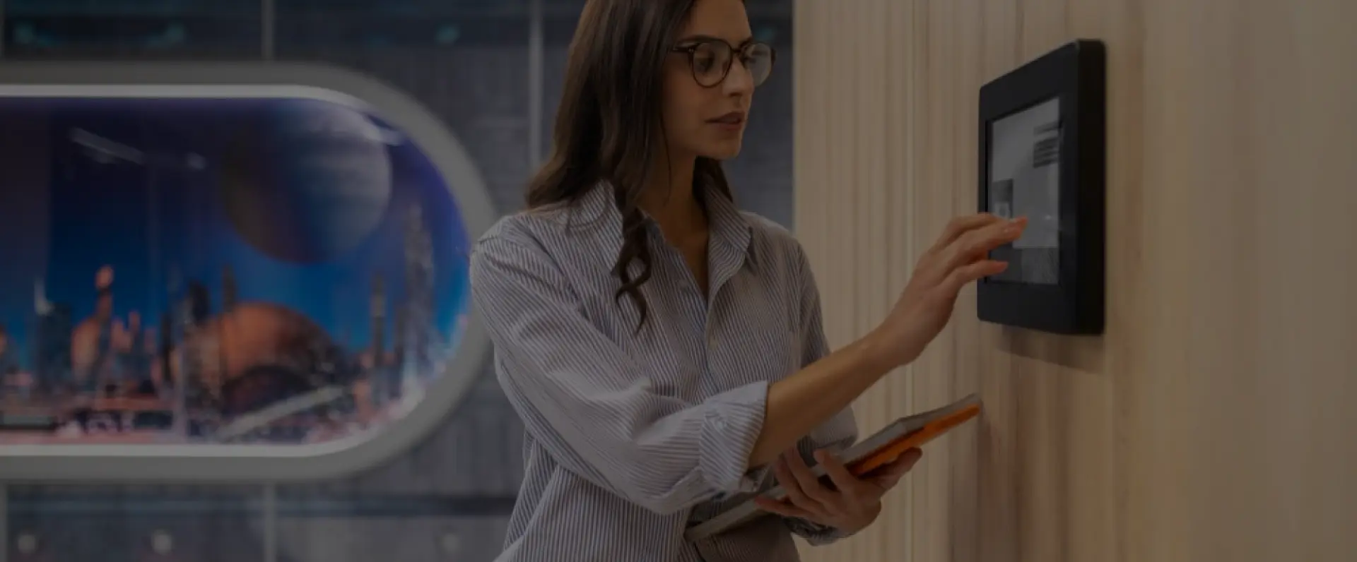 A woman in a striped shirt interacting with a touchscreen display, holding a tablet, in a modern interior setting.