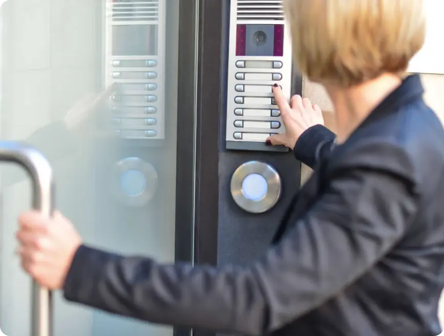 A person in a black blazer is pressing a button on a door intercom system.