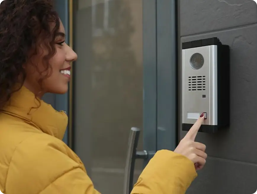 A woman in a yellow jacket smiling and pointing at a door intercom.