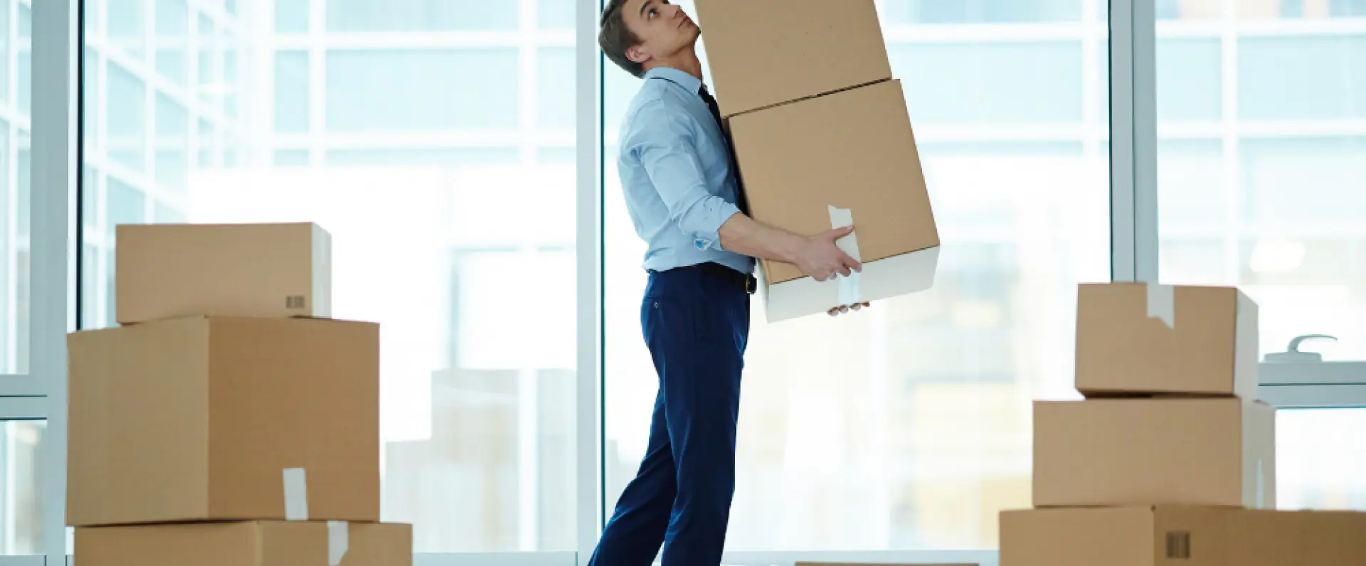 A man in a blue shirt and dark pants is lifting several boxes in an office setting with large windows.