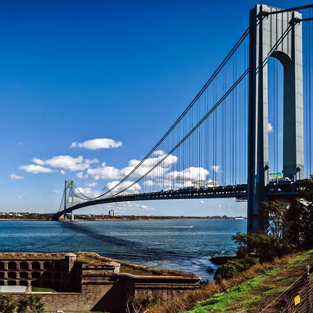 A tall suspension bridge with a blue sky and clouds in the background, over a body of water, with greenery in the foreground.