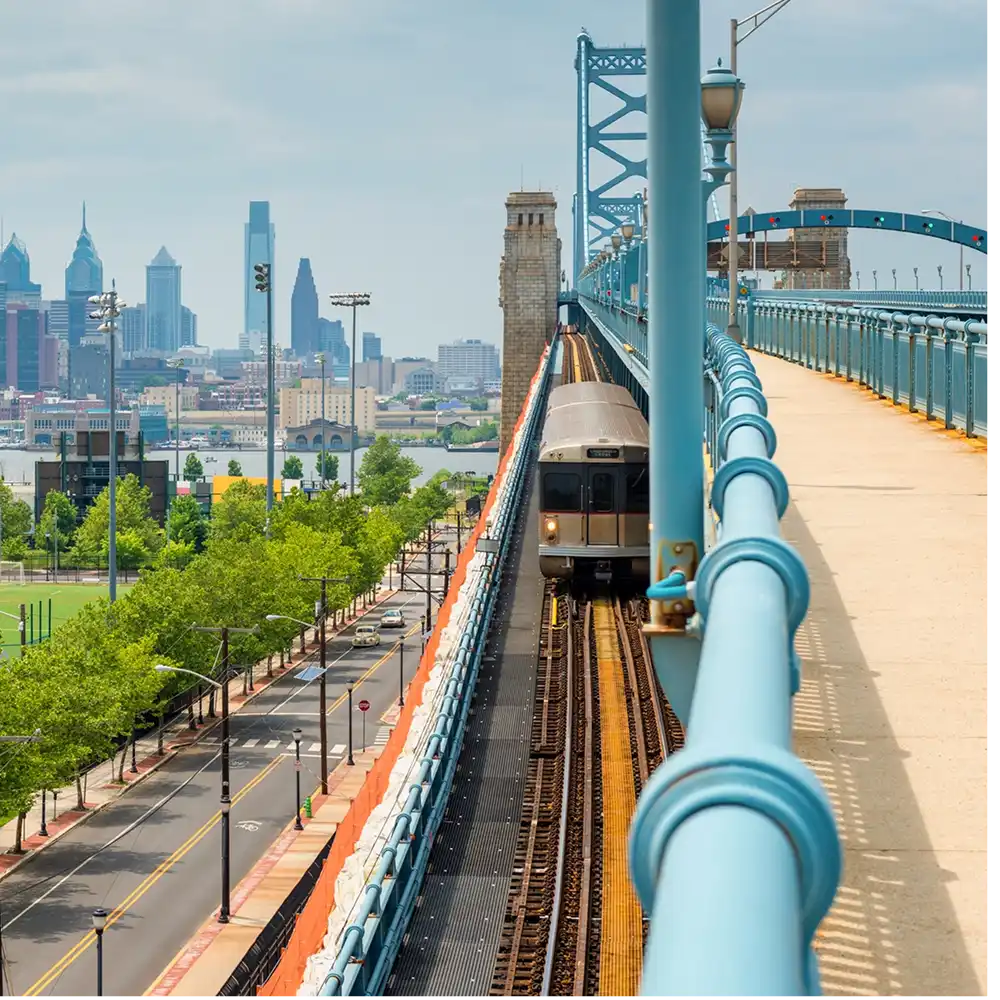 A train traveling on tracks with a city skyline in the background, featuring tall buildings and a bridge.