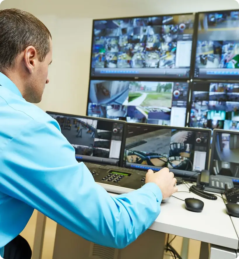 A man in a blue shirt monitoring multiple security camera feeds on large screens in a control room.