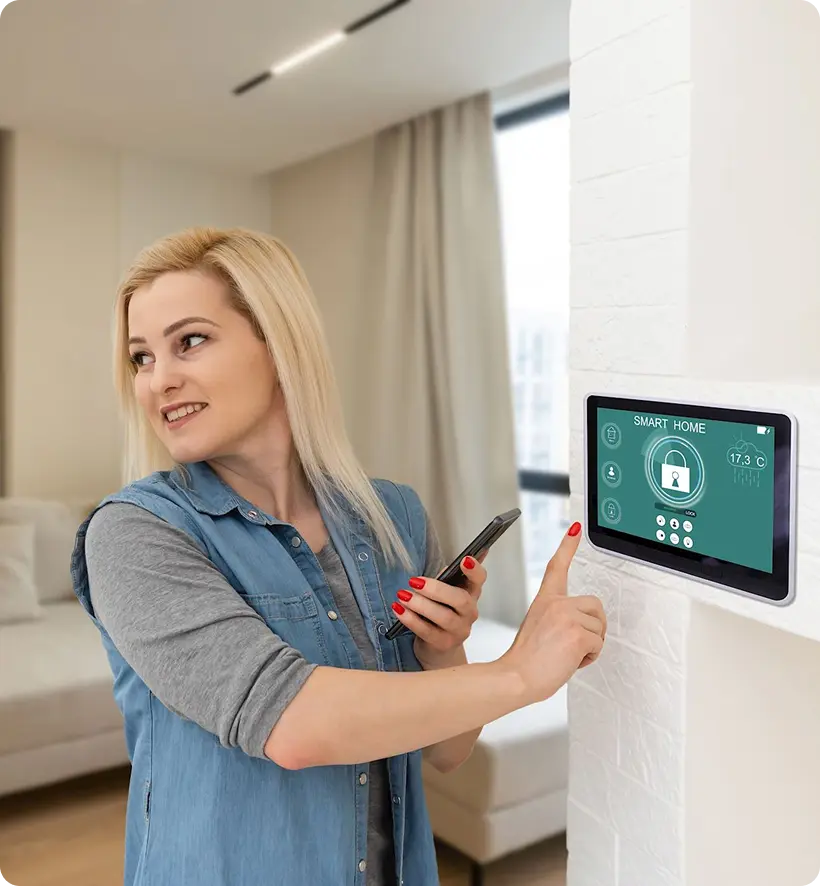 A woman with blonde hair smiles while using a smartphone and adjusting a smart home control panel on the wall.