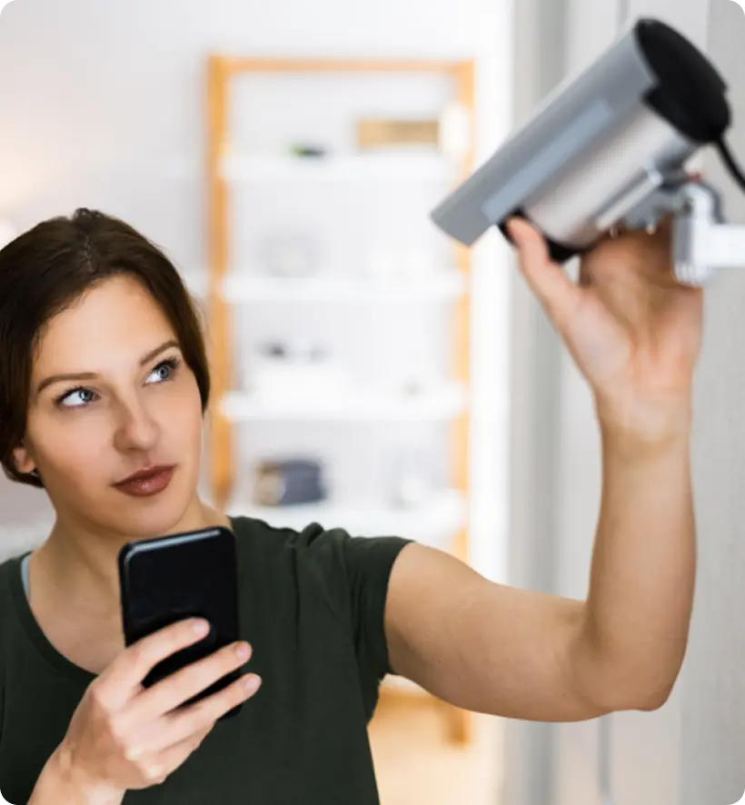 A woman installing a security camera while holding a smartphone.