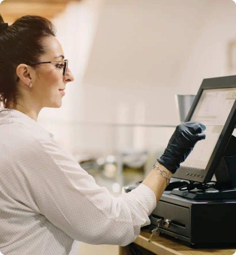 A woman with glasses and gloves operates a touchscreen point-of-sale system, standing in a bright indoor space.