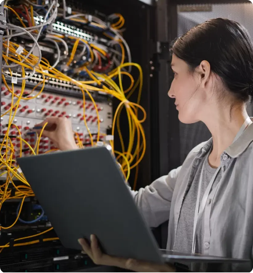 A woman working in a server room, connecting cables and holding a laptop.