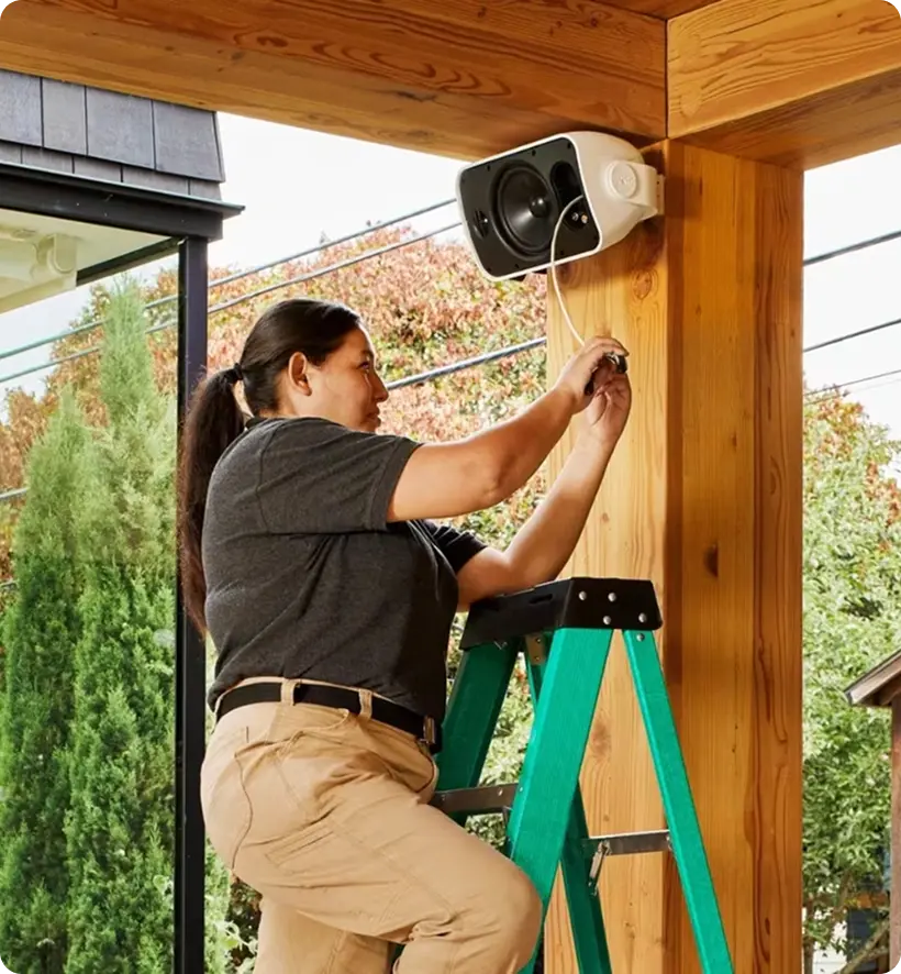 A woman is using a ladder to install a speaker on a wooden post.
