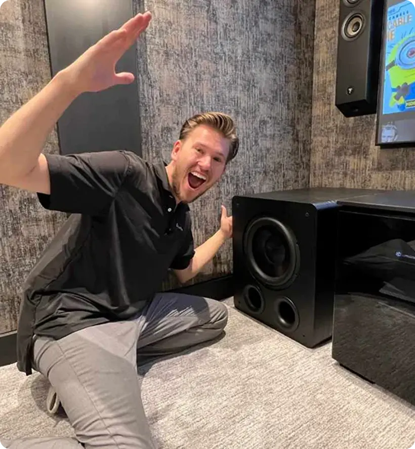 A man smiling and posing with a large speaker or subwoofer in a modern room with textured walls.