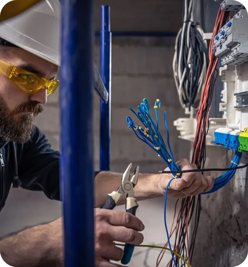 A worker in a hard hat and protective glasses is using pliers to work on electrical wiring connected to a circuit panel.