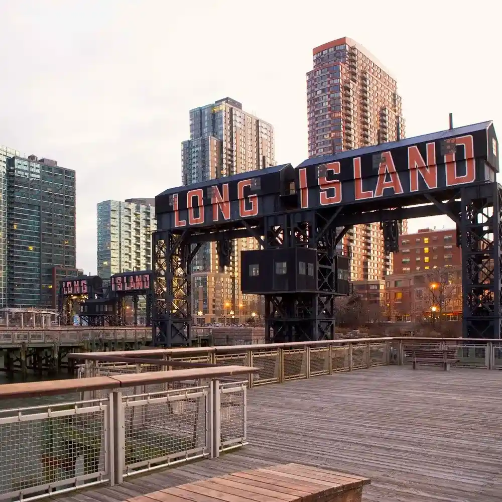 The image shows a waterfront area with a prominent sign that says "LONG ISLAND," surrounded by modern buildings and a wooden walkway or pier.